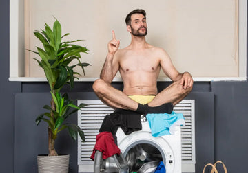 Lifestyle photo of a man sitting on a washing machine while wearing yellow briefs and pointing upward