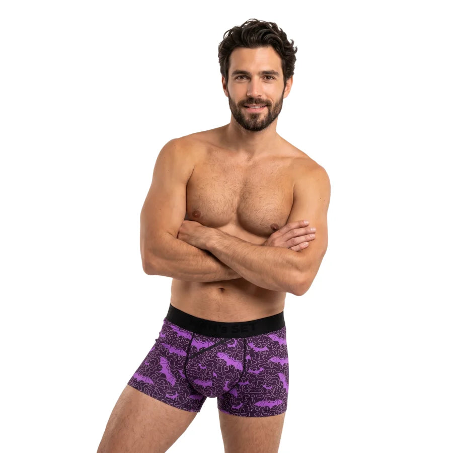 A shirtless man with a beard modeling purple and black printed trunks against a plain white background.