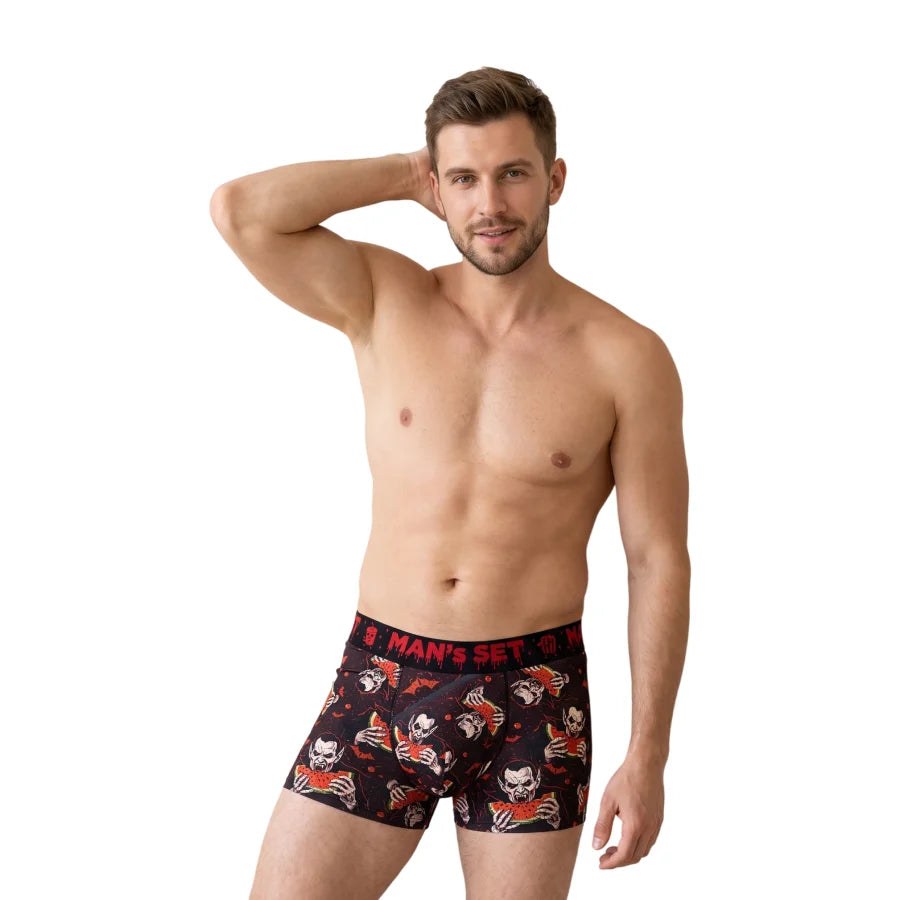 A shirtless male model wearing black boxer briefs with a red and white skull-and-crossbones pattern and a red waistband, posed against a plain white studio background.